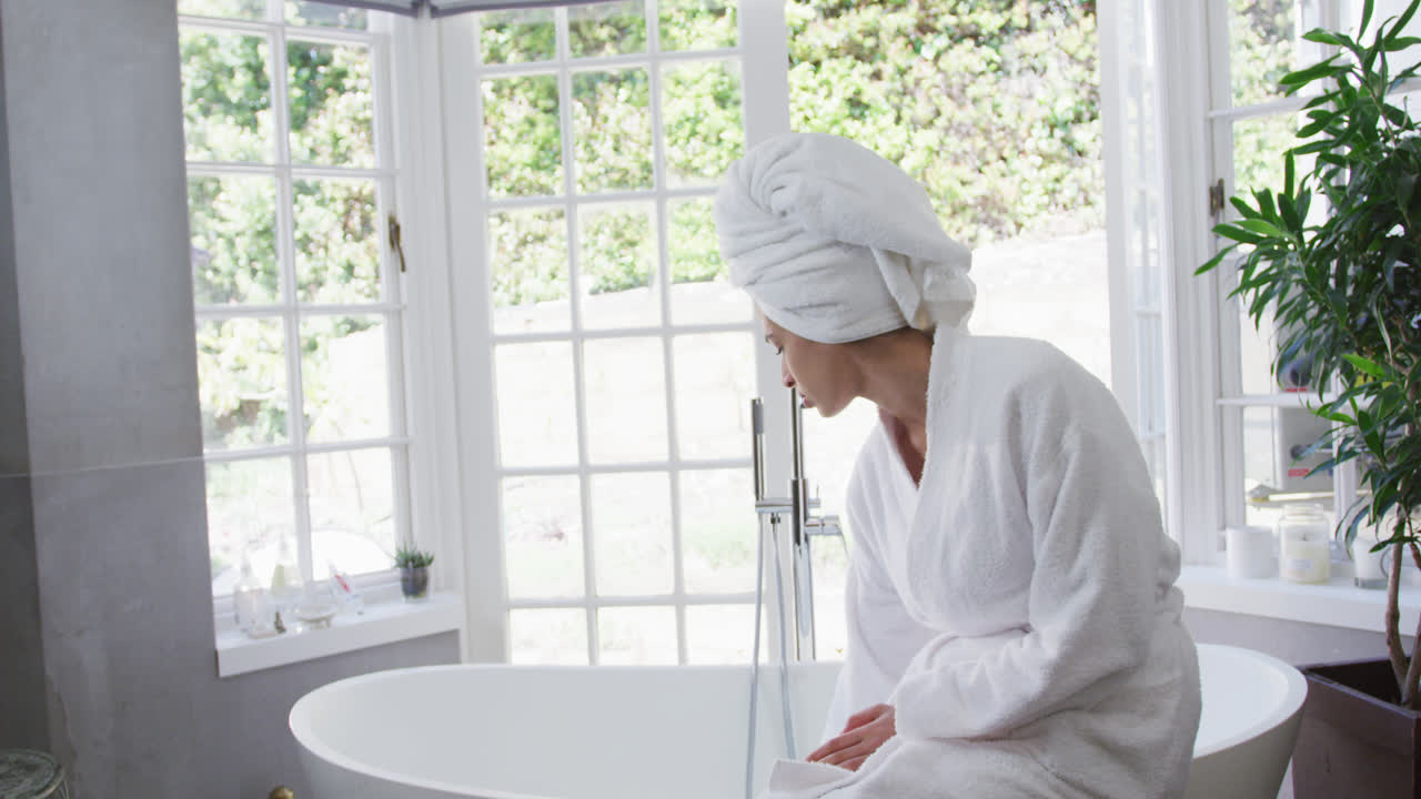 혼혈 여성이 물을 확인하고 욕실에서 욕조에 앉아 있습니다.
