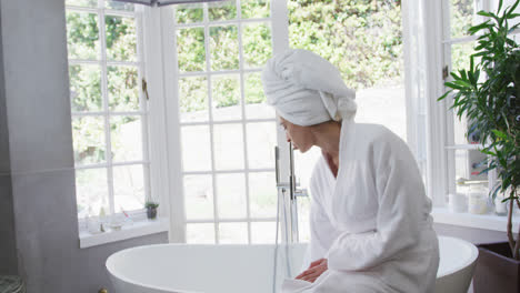 Mixed-race-woman-checking-water-and-sitting-on-bathtub-in-bathroom