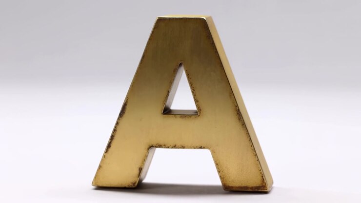 Scena in primo piano in movimento che mostra una lettera tridimensionale autoportante rifinita con vernice oro caldo con usura visibile sui bordi, patina brunastra e leggeri graffi concentrati attorno ai ritagli interni e alla base.