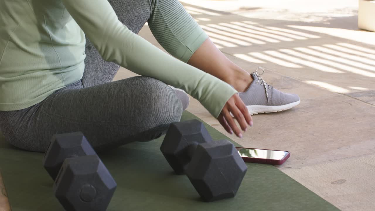 Sol brillando en el patio, una mujer levantando su smartphone y colocando una botella junto a unas mancuernas mientras se prepara para entrenar