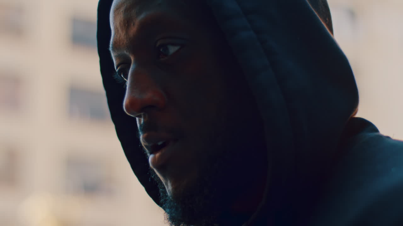 New York, USA, 8 April 2026: Close up portrait of man in black hoodie looking down. Tight headshot of Black man in hooded sweater with soft outdoor lighting and blurred architecture in background