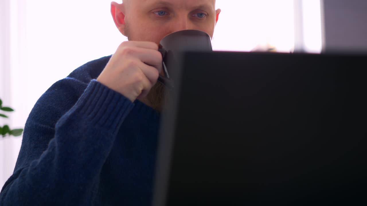 집에서 컴퓨터 앞에서 일하고, 아침 커피를 마시고, 노트북의 모니터 스크린 앞에서 수염을 가진 머리 남자가 근접합니다.