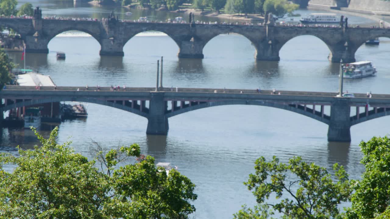 Daytime cityscape with boats moving under iconic Prague bridges, lush greenery, and calm river reflections
