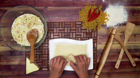 top view of rolling the spring rolls, marinated macaroni in the bowl, raw macaroni, wooden spoon, wooden spatula, flour, rolling pin, and chili on the table