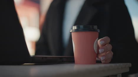 close-up of laptop on table with hand in black coat, polished nails, dropping a pink coffee cup onto table surface sits down, blurred background