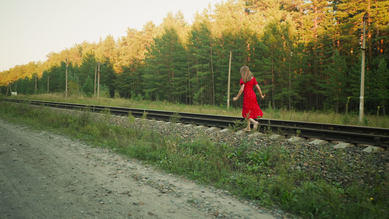 Rear view of young lady in red dress carefully jumping between railway sleepers along track in rural forest setting during golden hour, surrounded by trees, gravel, poles, and soft natural sunlight