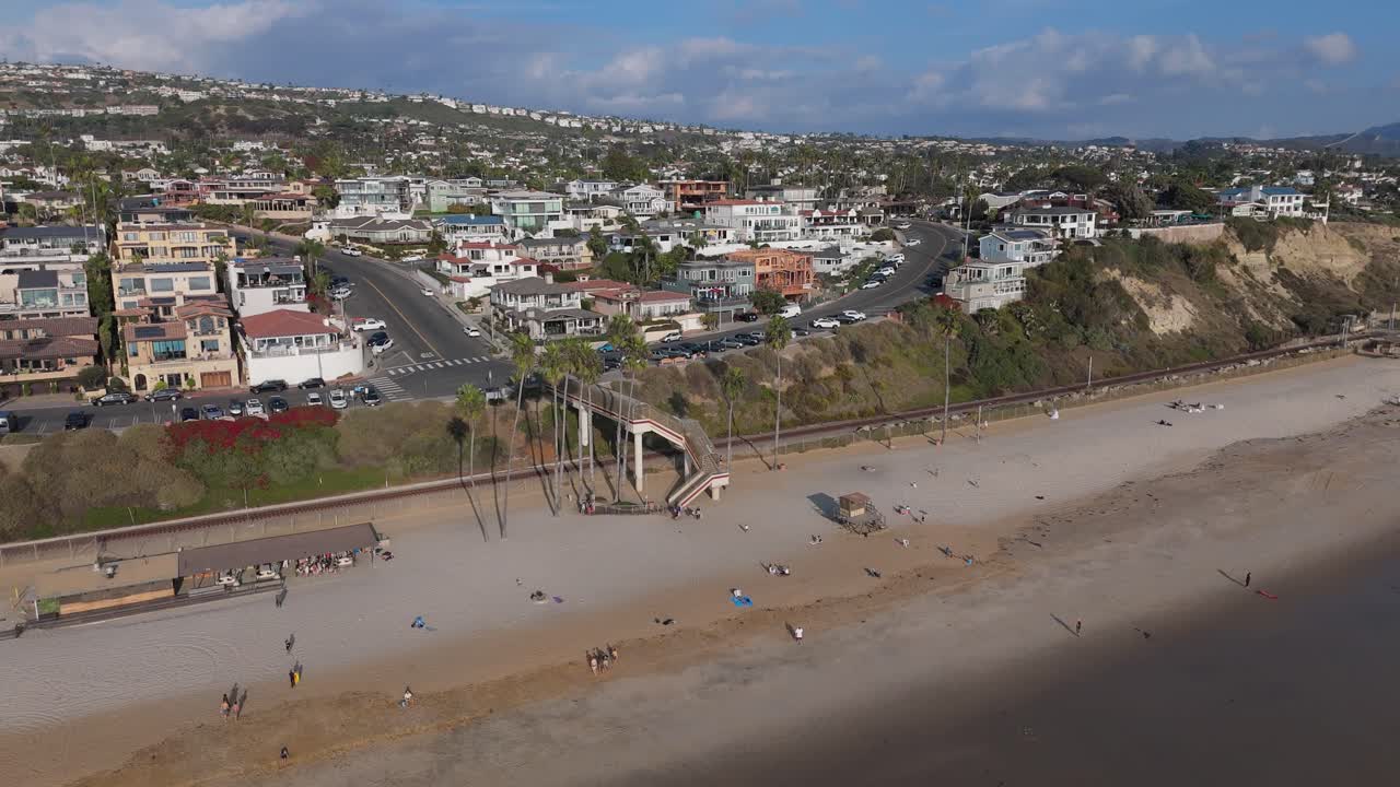 A wide drone-captured aerial view of the T-Street bridge spanning over railroad tracks, with scenic views of beachfront homes and coastline in San Clemente, California, USA.