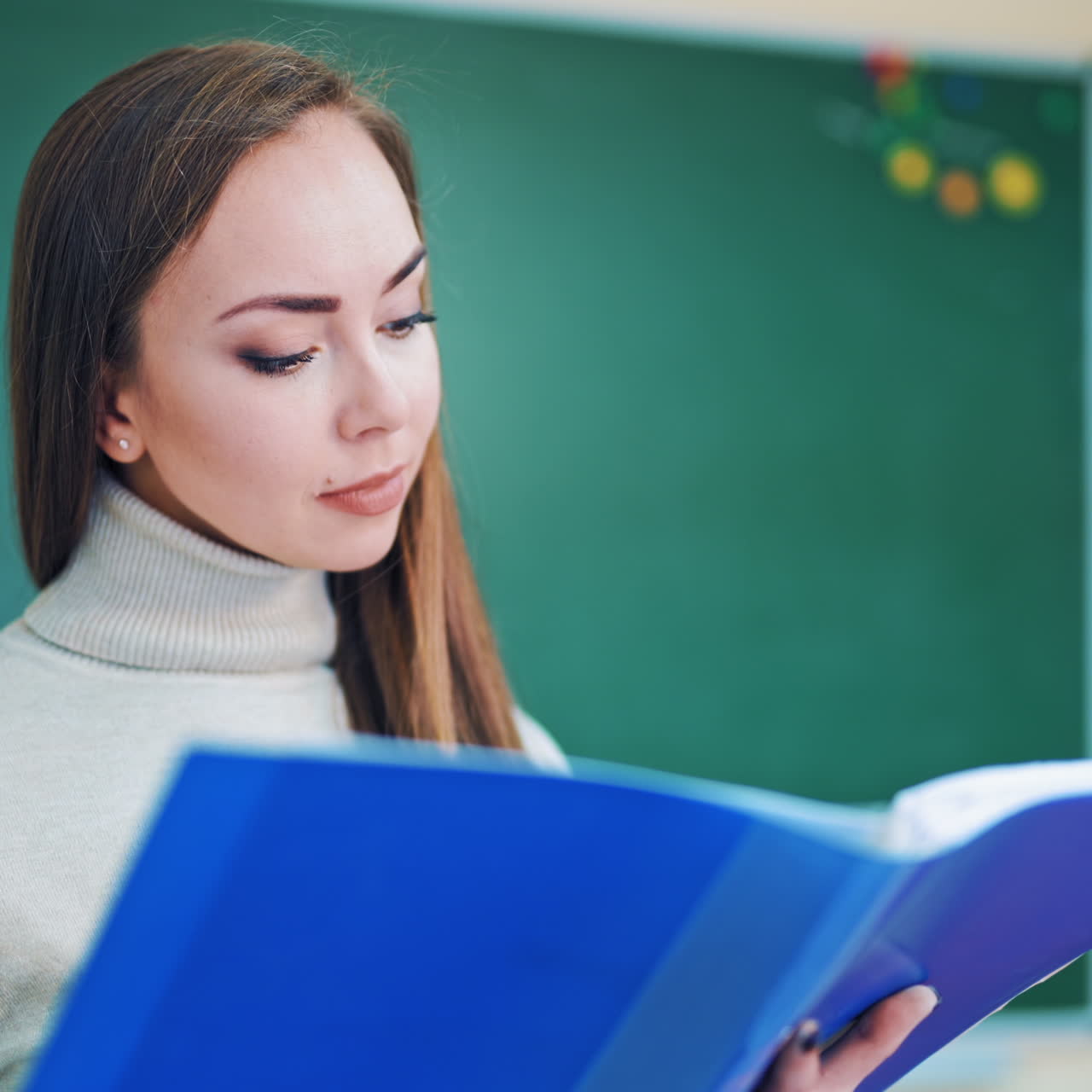 Portrait of a beautiful teacher. Lovely face of a young woman looking into a blue folder on the school board background. Close-up.