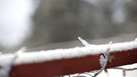 Nahaufnahme-Der-Weißen-Weihnachtsbeleuchtung-Auf-Einer-Verschneiten-Terrasse-Mit-Pinien-Im-Hintergrund-Und-Schneefall