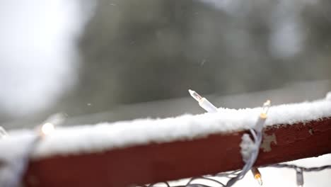 Close-up-of-white-Christmas-lights-on-a-snowy-deck-with-pine-trees-in-the-background-and-snow-falling