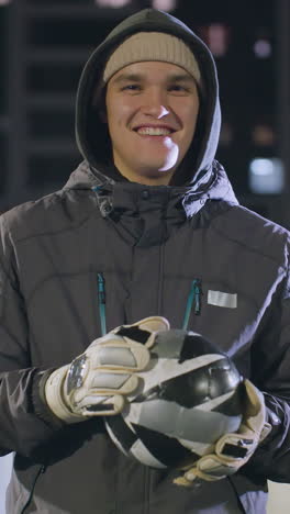 atleta sonriente con capucha y guantes sosteniendo una pelota de fútbol en un campo deportivo urbano por la noche, edificios de gran altura iluminados, postes de portería y luces bokeh vibrantes
