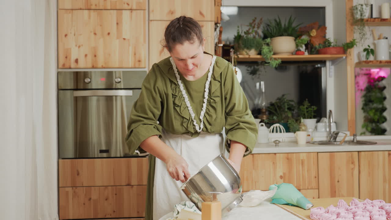 Focused caterer in green outfit and white apron uses wooden spatula to scrape dough from metal bowl into piping bag while preparing dessert in warm rustic kitchen with plants, oven, and baking tools