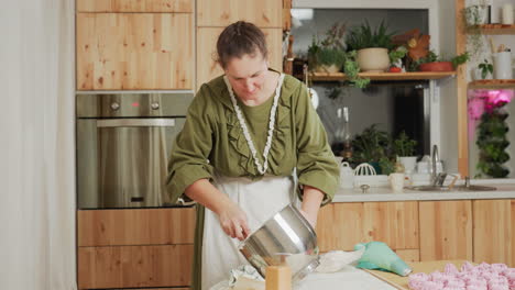 focused caterer in green outfit and white apron uses wooden spatula to scrape dough from metal bowl into piping bag while preparing dessert in warm rustic kitchen with plants, oven, and baking tools
