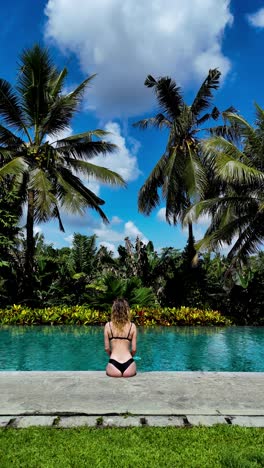 vertical drone pull-back reveals a blonde woman in bikini resting at the edge of a luxury villa pool amid lush palms and tropical jungle near ubud, bali, on a bright sunny day