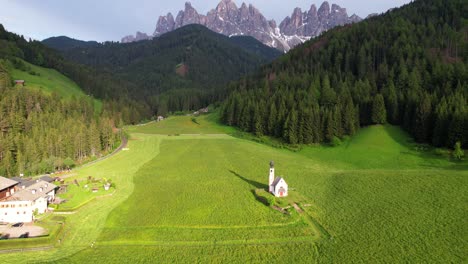 Aerial-tilt-up-of-San-Giovanni-in-Ranui-Church-revealing-Odle-mountain-range,-Italy