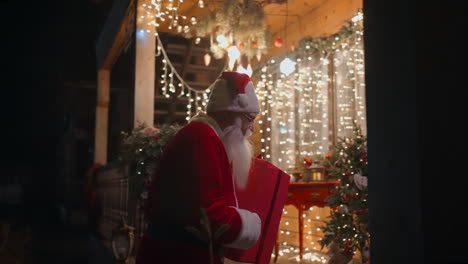 santa claus carries the gift in his hands and enters the children's house to leave gifts under the christmas tree. decorated christmas house