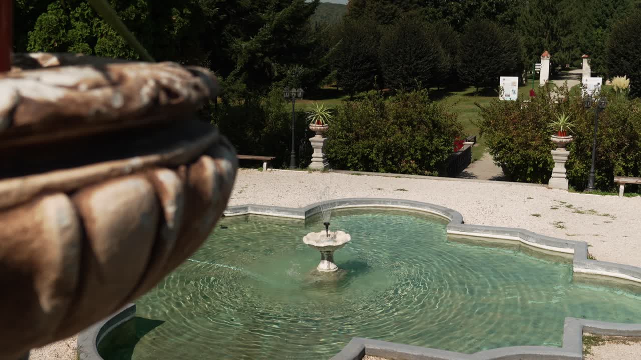 Wide shot of a stone water fountain surrounded by greenery in a peaceful garden under bright summer sunlight