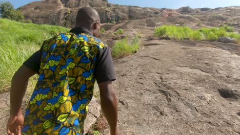 tracking shot from behind of an african man climbing up a steep granite rock in the hot african sun while wearing a colorful african shirt