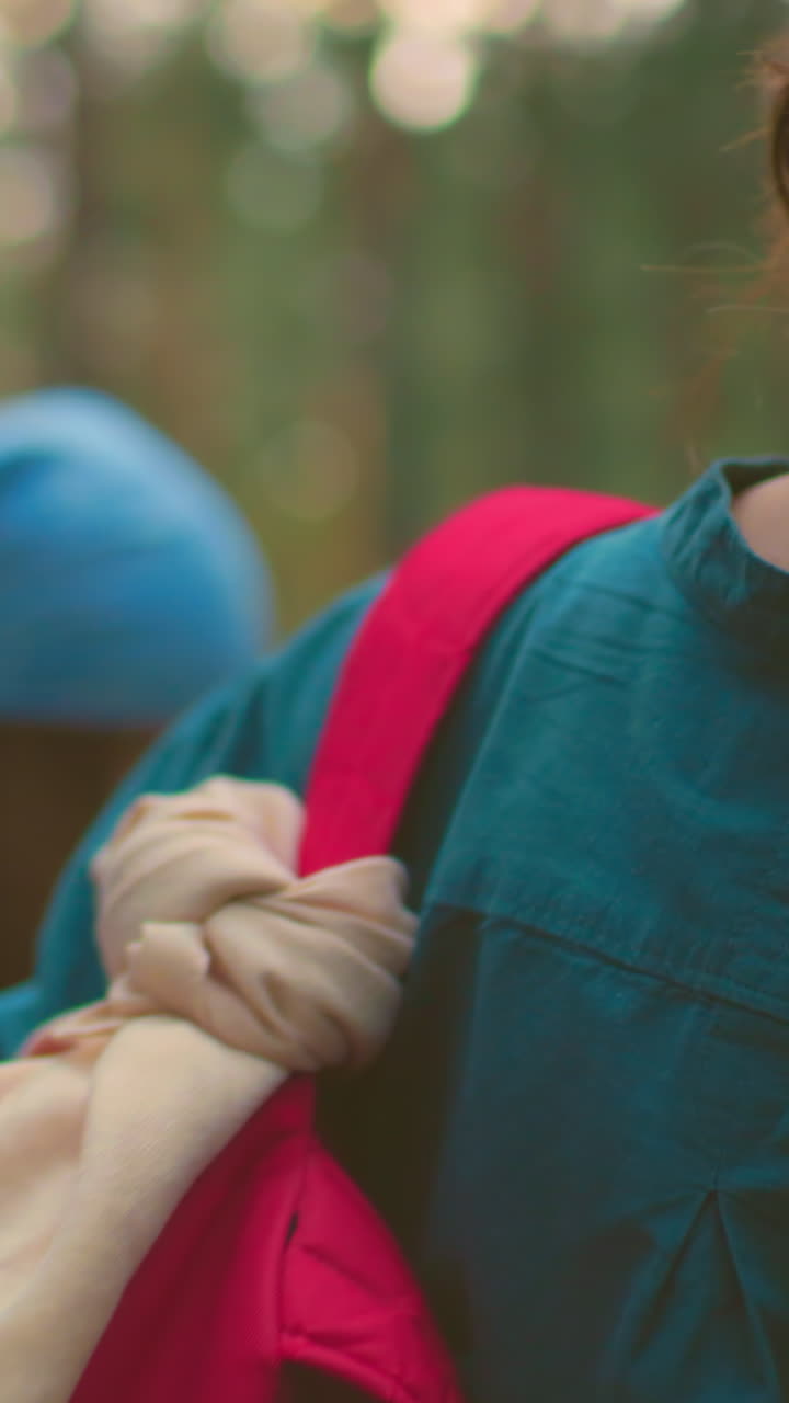 vista trasera de una mujer en el bosque con una camisa azul azul, colgando una bolsa roja con un envoltorio de tela sobre el hombro, el cabello atado en un pan, se aleja con su amiga borrosa en el fondo con vegetación