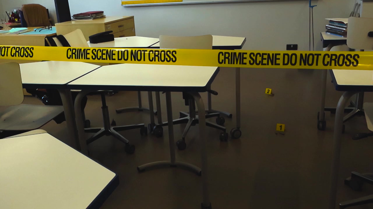 Nastro giallo della polizia con la scritta scena del crimine non oltrepassare che delimita un'aula vuota con indicatori di prove sul pavimento, suggerendo un evento violento come una sparatoria scolastica o un omicidio