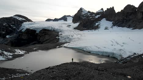 aerial flyover over a hiker overlooking the glacial lake and ice of the claridenfirn glacier in uri, swizerland with a view of the alpine peaks and icebergs floating in the water during sunset