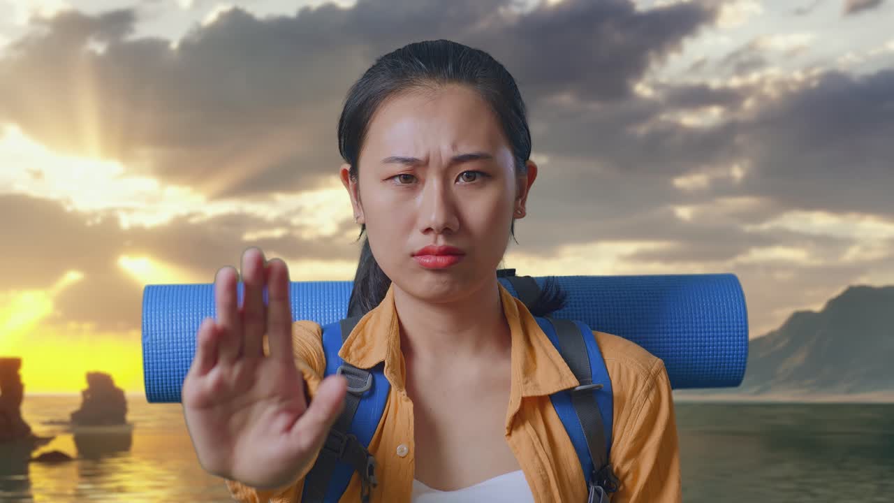 Close Up Of Asian Female Hiker With Mountaineering Backpack Looking At Camera And Making Stop Hand Sign While Standing on Beach During Sunset Time