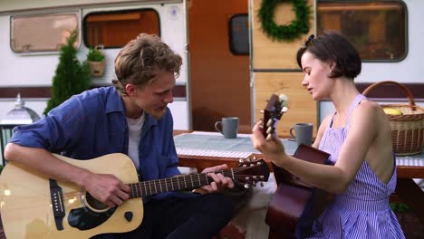 bearded guy teaching her girlfriend play the guitar outdoors sitting close to their trailer house. holding acoustic guitars, man showing how to tap particular chords. close up