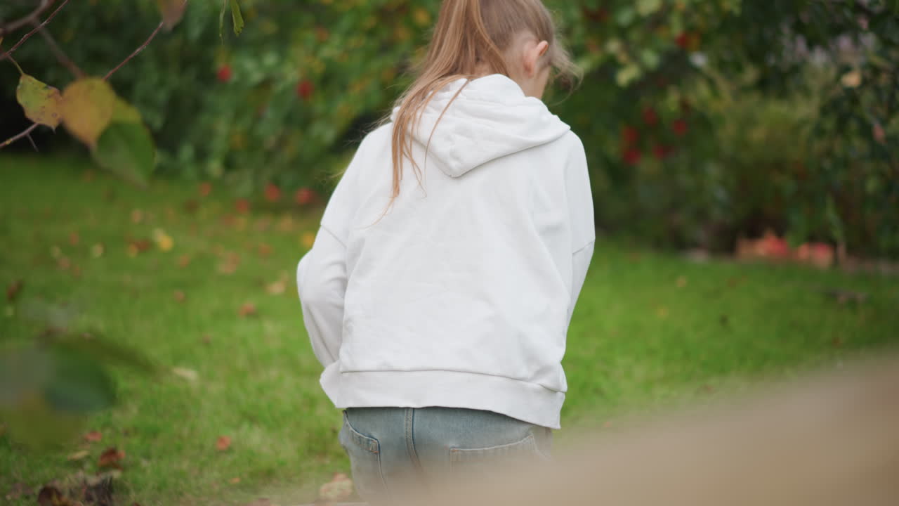 Weißes Mädchen mit Kapuzenpullover beugt sich über das Gras. Eine ruhige Hinterhofgarten-Szene, in der gefallene Blätter gesammelt werden. Pferdeschwanzzopf. Weiches Herbstlicht, nachdenkliche Stimmung. Nahaufnahme, natürliche Texturen für Archivmaterial.