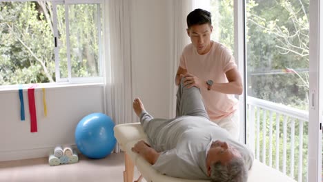 diverse male physiotherapist advising and senior male patient exercising on couch, in slow motion