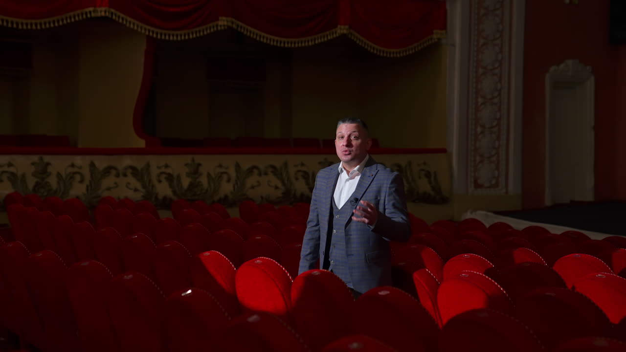 Monologue of an actor in empty theater. Professional performer in suit standing among red chairs in theatrical hall and talking on camera. Pandemic.