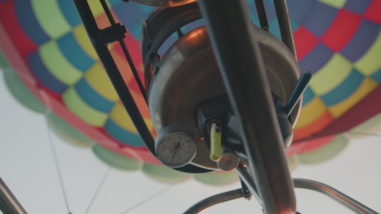 Close up of gloved hand gripping control valve of metal burner beneath vibrant hot air balloon canopy, with pressure gauge visible, capturing technical detail of balloon flight setup