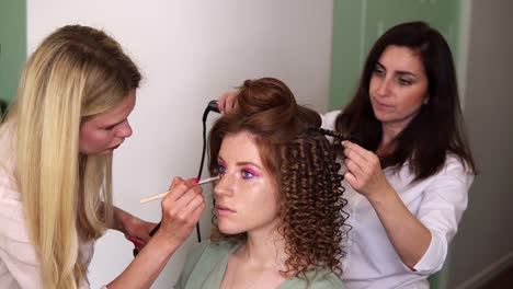 Two-artists-working-with-client-in-tandem---makeup-artist-applies-toner-on-the-face-of-a-beautiful-model-with-freckles,-red-headed.-Hairdresser-with-a-curling-iron-making-hair-style.-Slow-motion