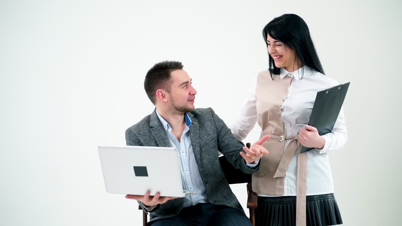 Businessman talking to a young smiling secretary. Handsome man in suit sitting with a laptop in his hands and talking to a beautiful woman with a folder.