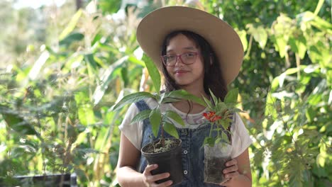 Una-Chica-Sonriente-Con-Sombrero-Y-Gafas,-Parada-En-Medio-De-Un-Jardín-Botánico-Con-Una-Planta-De-Aguacate-Y-Una-Maceta-De-Flores-En-Las-Manos.