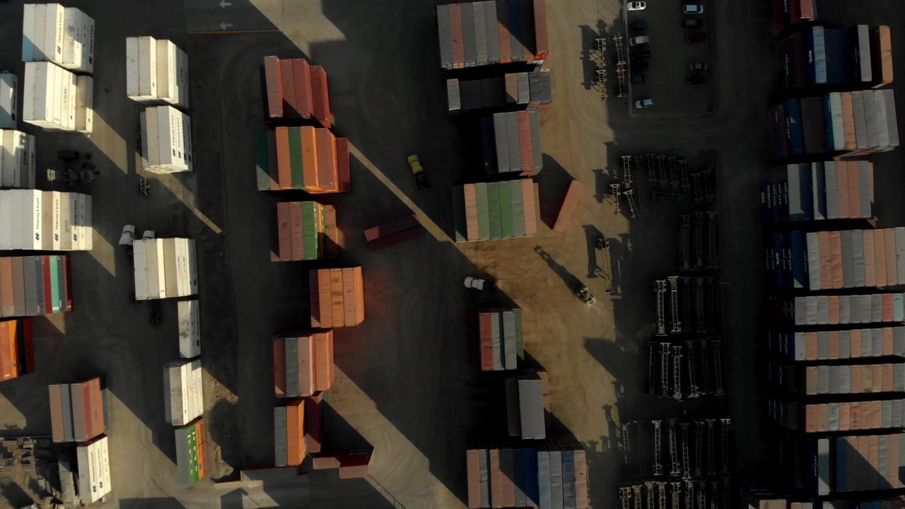 Aerial drone view of shipping containers arranged in rows across a paved cargo yard, showing colorful container units, parked vehicles, equipment, and long shadows.