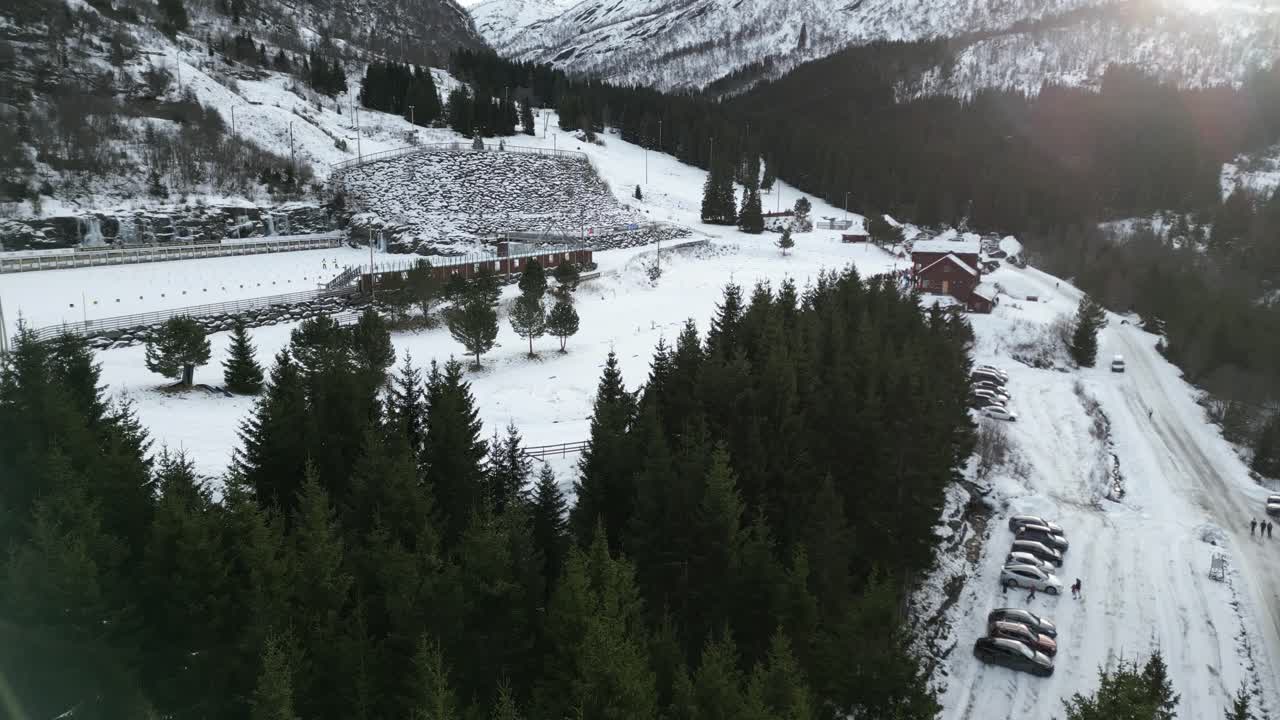 4K drone footage panning sideways over Vik Skisenter in Norway, showing skiers training on snowy tracks surrounded by pine forest and mountain scenery in winter sunlight