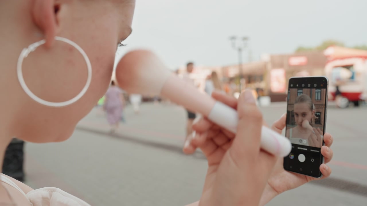 Young woman applies makeup with large brush while another woman records her on smartphone in outdoor public space. scene features soft natural light and blurred background activity focusing