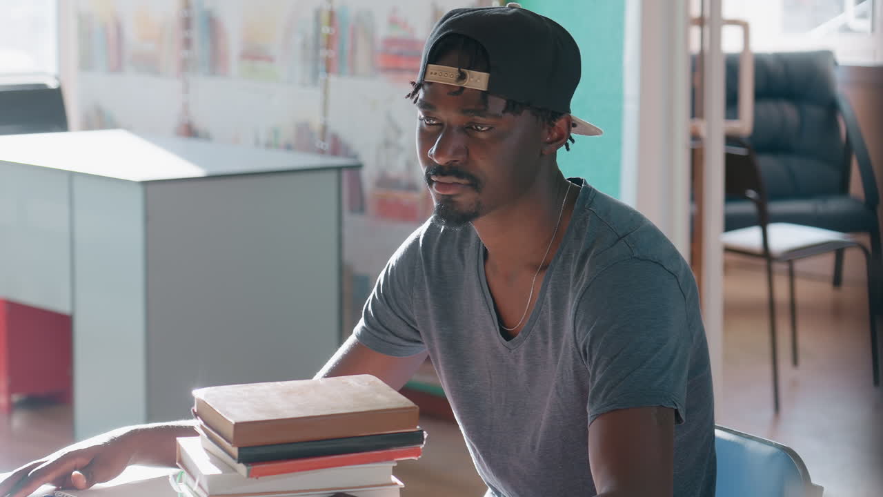 Calm young man in cap sits at library table beside stack of books, arms resting, gazing ahead in soft daylight, relaxed focus, modern study space with shelves and chair blurred in background