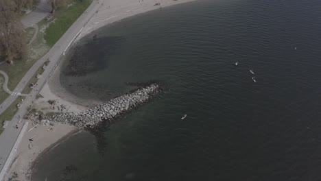 Aerial-drop-over-English-Bay-Sunset-Beach-birds-eye-view-of-paddle-board-surfers-enjoying-an-entertaining-summer-experience-of-physical-fitness-while-people-excercise-on-the-seawall