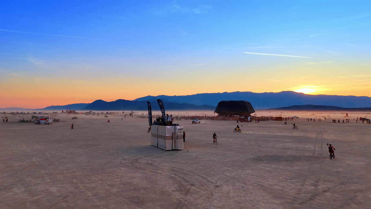 Desert platform scene in fading light. A raised viewing platform stands among cyclists and walkers as dusk settles across the expansive playa