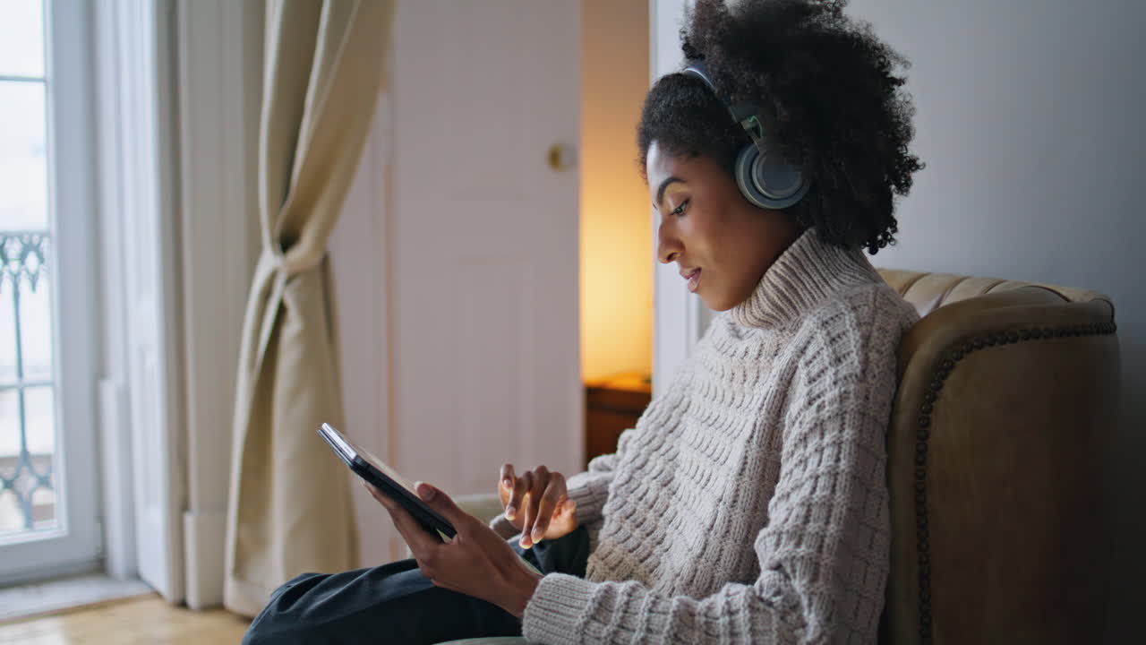 Focused girl scrolling pad comfy home. African headphones lady listening music