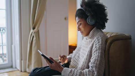 focused girl scrolling pad comfy home. african headphones lady listening music