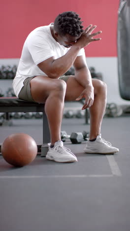 vertical video: resting on bench, strong and fit man wiping sweat in gym with basketball nearby