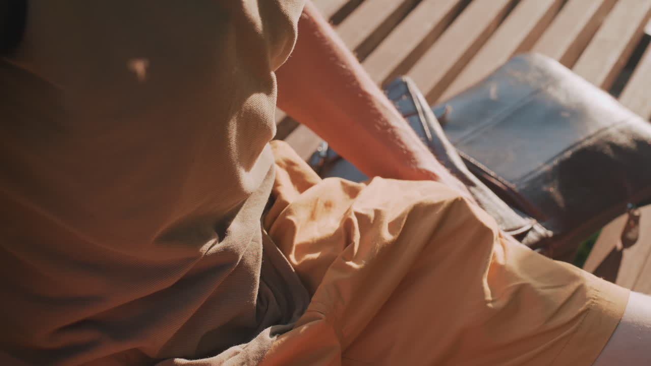 Uncomfortable Individual On Bench, Man Exhibiting Nervous Gestures While Resting In Sunlight Outdoors, Fidgeting Man Displaying Anxious Actions While Seated On Park Bench In Sunny Weather