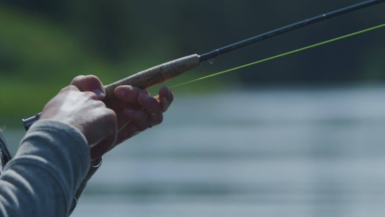 Rekaman gerak lambat jarak dekat seorang pria memancing dengan teknik fly fishing, dengan ikan di kailnya, senar terulur, dan dia menggulungnya kembali