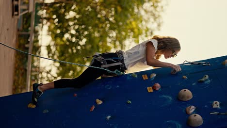 Vertical-video:-A-blonde-girl-in-a-white-T-shirt-in-special-equipment-and-folding-insurance-climbs-up-a-blue-climbing-wall-in-the-summer.-Blonde-girl-climber-trains-ska-climbing-and-climbs-up-a-blue-climbing-wall-in-summer