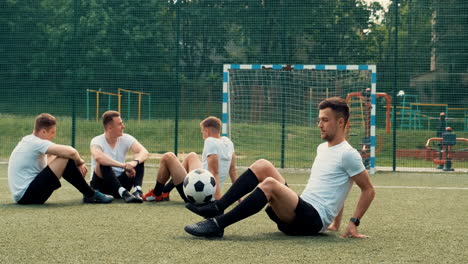 male street football player sitting on grass and bouncing soccer ball with feet while team resting on pitch