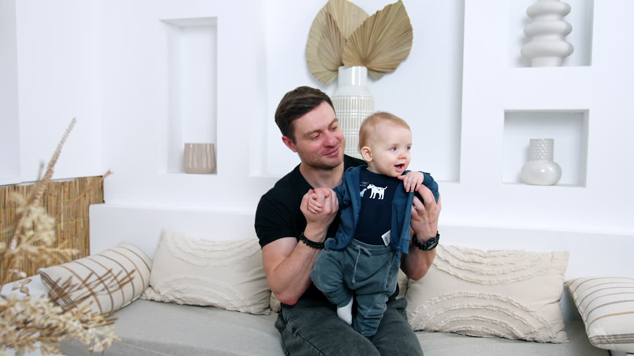 Man in black t-shirt sits on the sofa holding his son. Little kid smiles to camera, dad kisses and hugs his adorable baby.