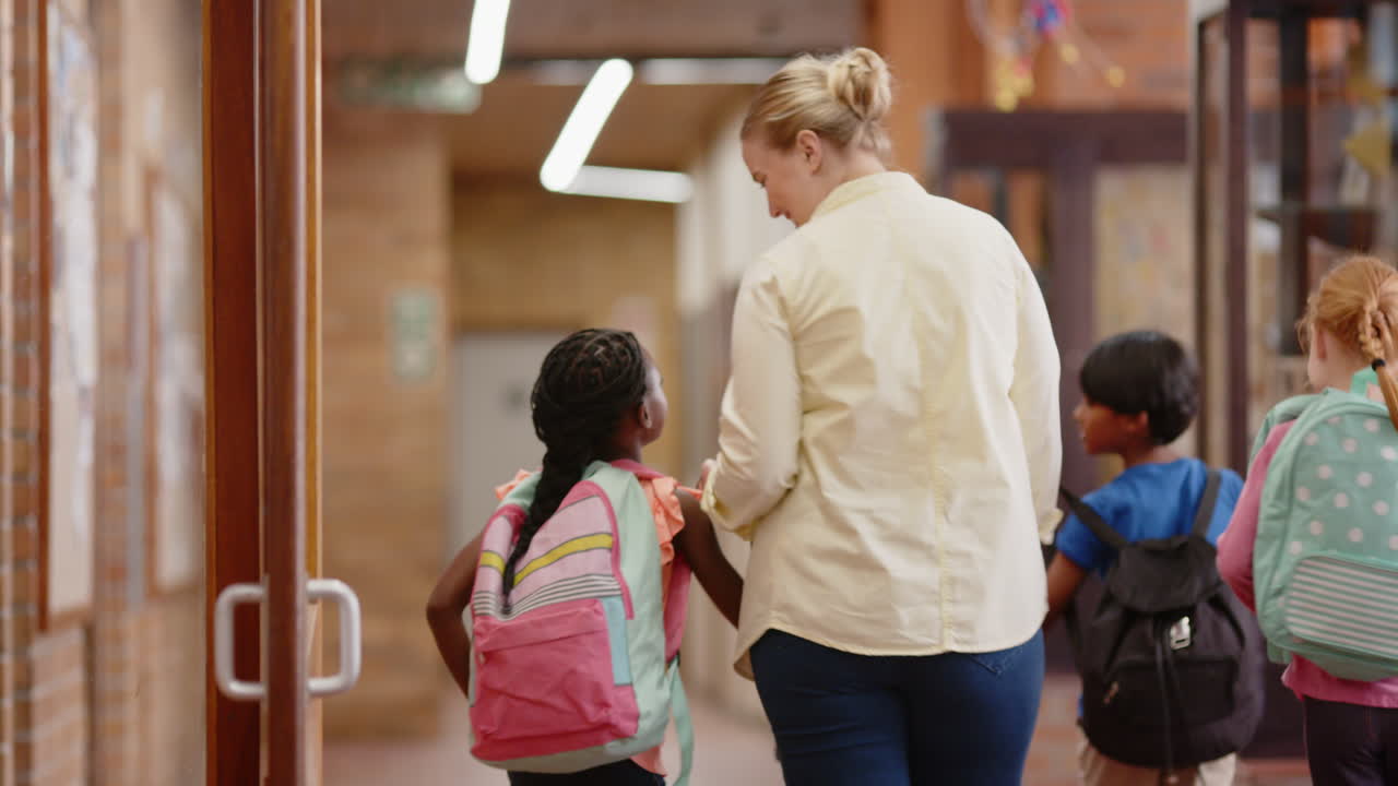 Walking in school hallway, female teacher holding hand of student with backpack
