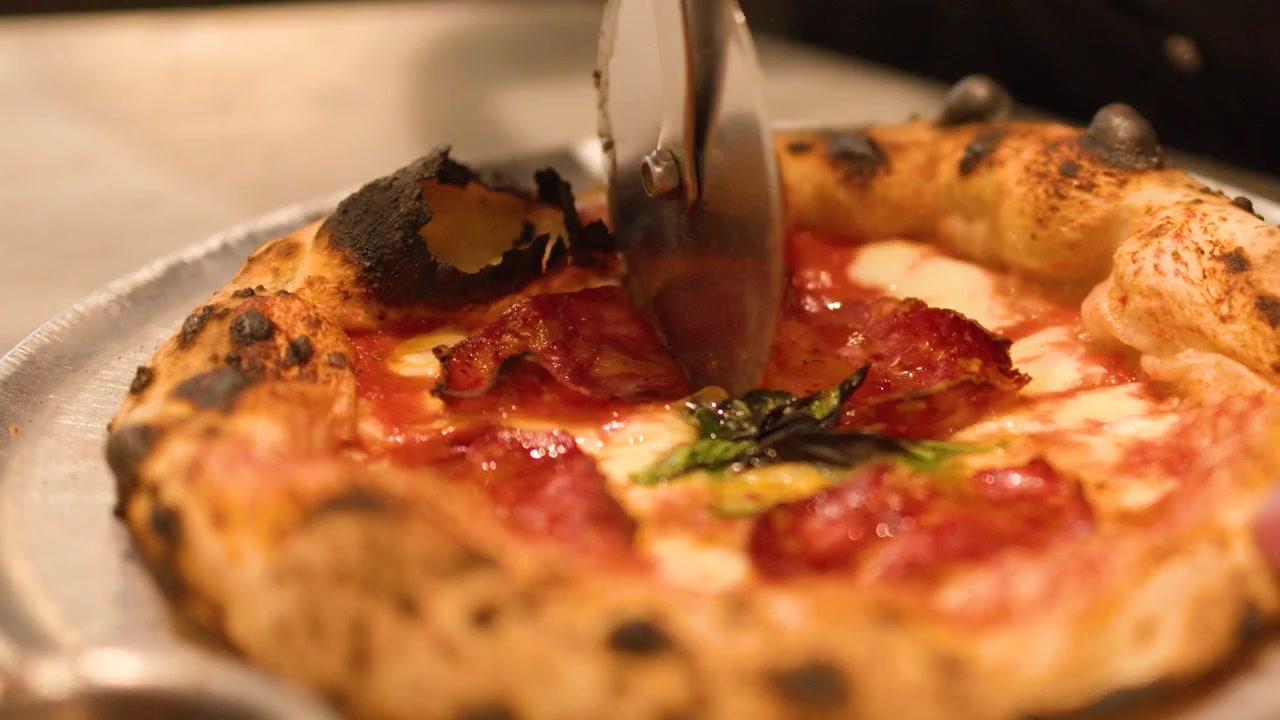 Primo piano di mani che tagliano una pizza napoletana calda con salame piccante e basilico su un vassoio.